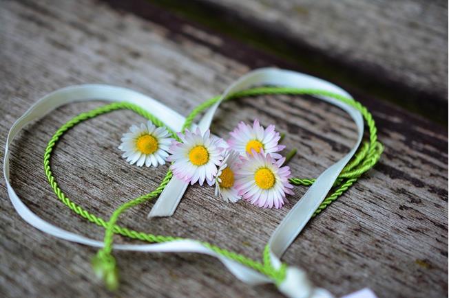 A heart made of green and white ribbon, decorated with small daisies, is placed on wood.